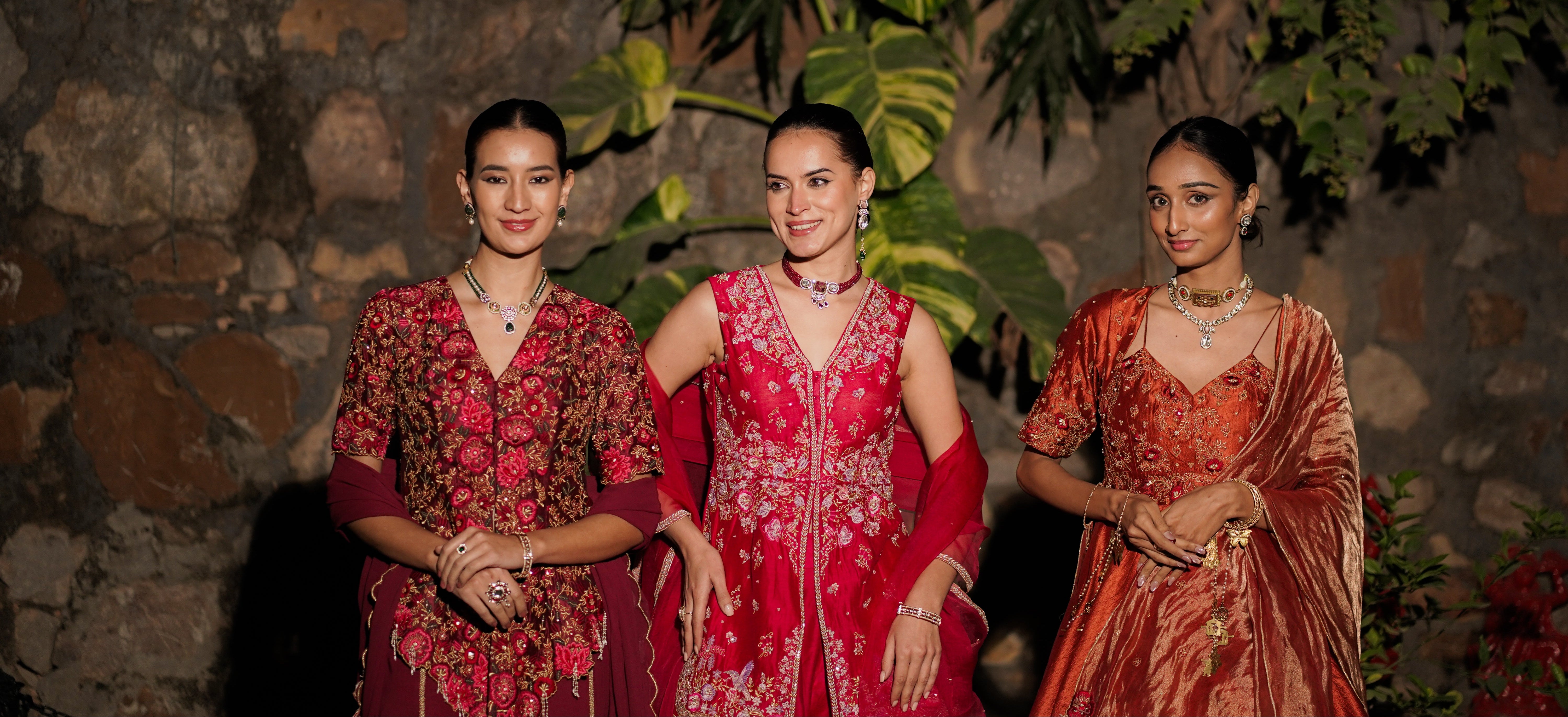 Three women in traditional red and patterned dresses standing against a stone wall with greenery.